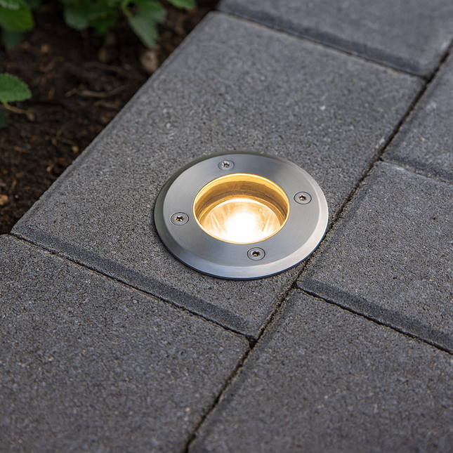 Round outdoor light fixture embedded in a paved pathway with greenery in the background.