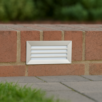 White rectangular vent on a red brick wall with blurred greenery in the background