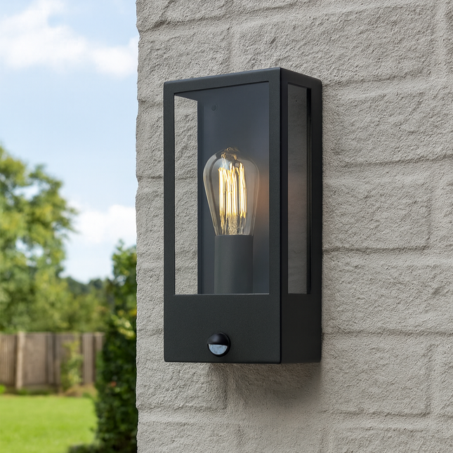 Black outdoor wall light fixture on a stone wall with a garden and sky in the background